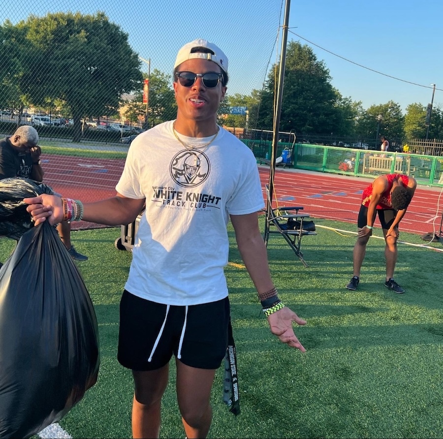Isaiah Kirby outdoors on a running track, wearing sunglasses and a White Knight Track Club shirt